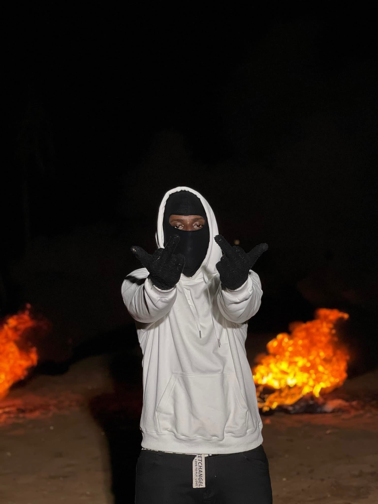 Portrait de nuit de Le Mélangeur en hoodie blanc et cagoule noire, devant des flammes