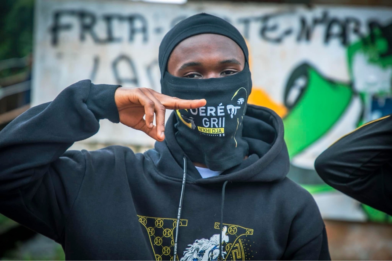 Portrait de Le Mélangeur en cagoule noire devant un graffiti « FRIA »